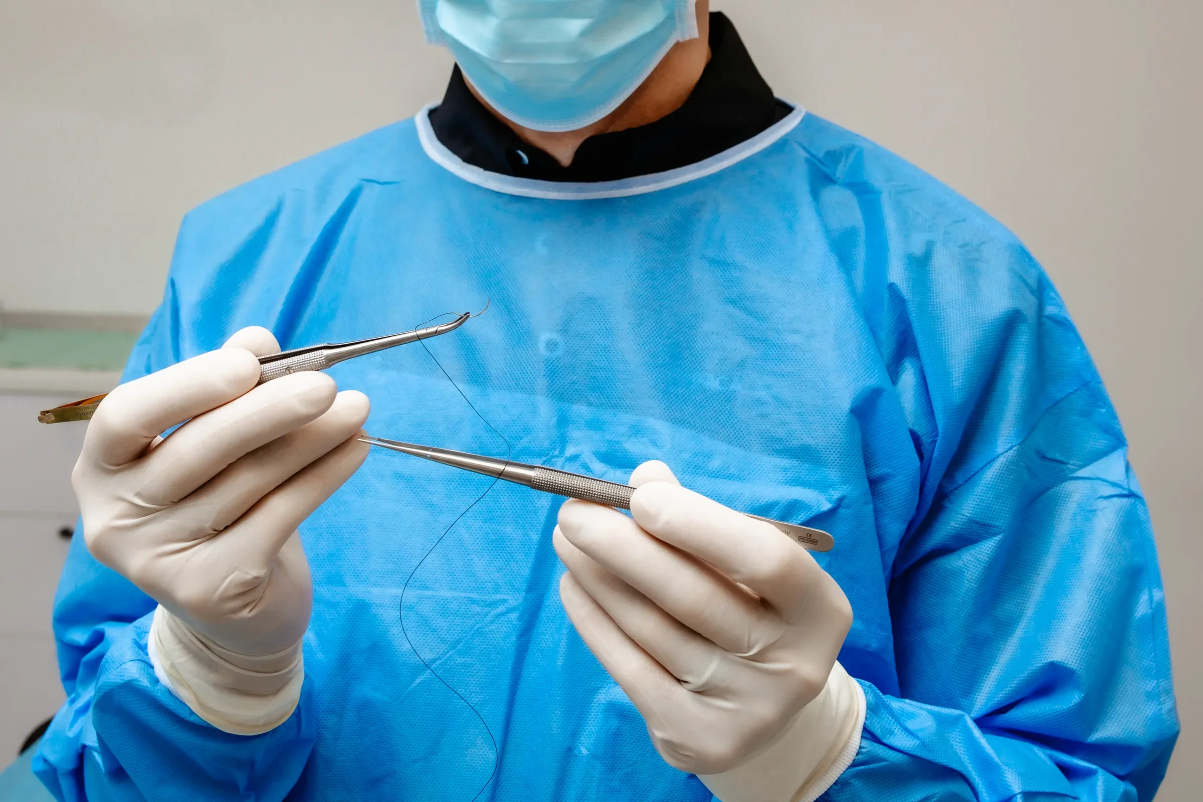 Mâinile medicului în mănuși, cu portacul, firul de sutură și pensa